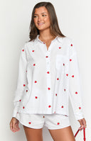 Model is wearing a white long-sleeve button-down shirt adorned with small red embroidered strawberries all over. The shirt is partially unbuttoned and paired with matching white shorts that also feature strawberry embroidery and a visible 