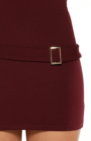 A lifestyle shot of our model wearing a form-fitting burgundy sleeveless mini dress. This mini dress features a high boat neckline and a decorative tonal belt at the hip accented with a rectangular gold buckle and a hanging fabric tab.