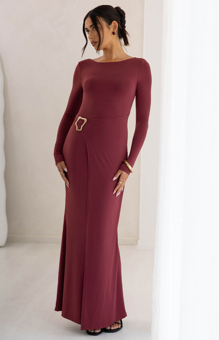 Full length view of a burgundy long sleeve maxi dress with a boat neck and a gold abstract buckle at the wrap waist