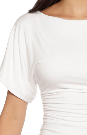 A lifestyle shot of our model wearing a short-sleeved white top. This garment features a wide boatneck fluttery cap sleeves and horizontal ruched gathering along the sides of the bodice.
