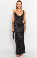 Blonde girl in black satin ruffle maxi dress and nude heels standing at a bar.