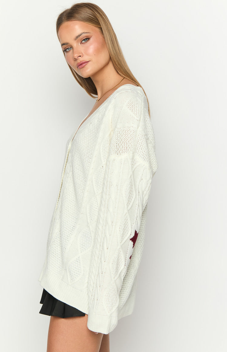 Side view of an off-white cable knit cardigan with a deep V-neck, intricate cable pattern, and a red star on the left sleeve