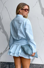 Rear view of a light blue long sleeve mini shirt dress with a cinched waist and tiered ruffled skirt