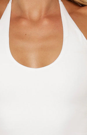 A studio shot from the neck to the waist of a woman wearing a plain white halter-style top. The garment features a scoop neckline and wide straps that tie behind the neck. It has a form-fitting silhouette with slight gathering at the side hems.