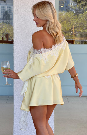 Side view of a yellow satin off-shoulder mini dress with white lace trim, loose sleeves, and hanging side lace detail