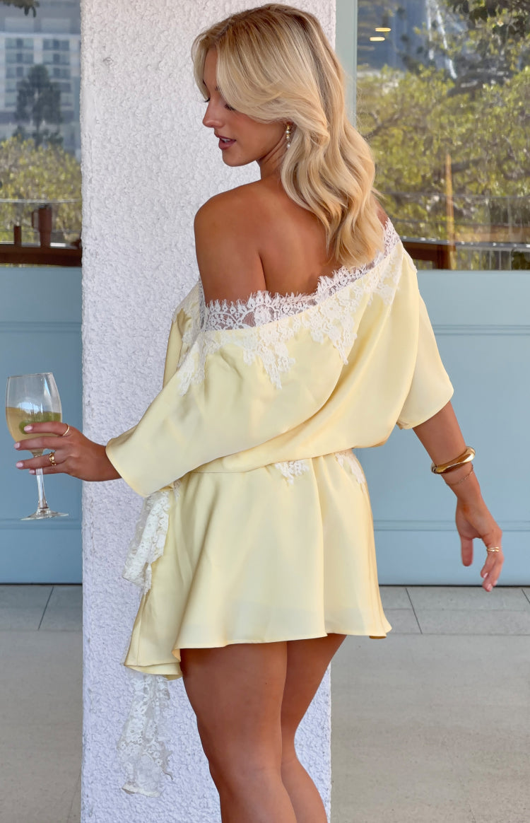 Side view of a yellow satin off-shoulder mini dress with white lace trim, loose sleeves, and hanging side lace detail