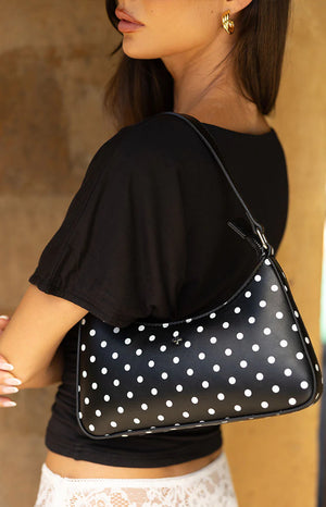 A lifestyle shot of our model carrying a small structured shoulder bag over her right arm. This bag is made of black material with a white polka-dot pattern and features a black strap and a small silver cross-shaped emblem on the side.