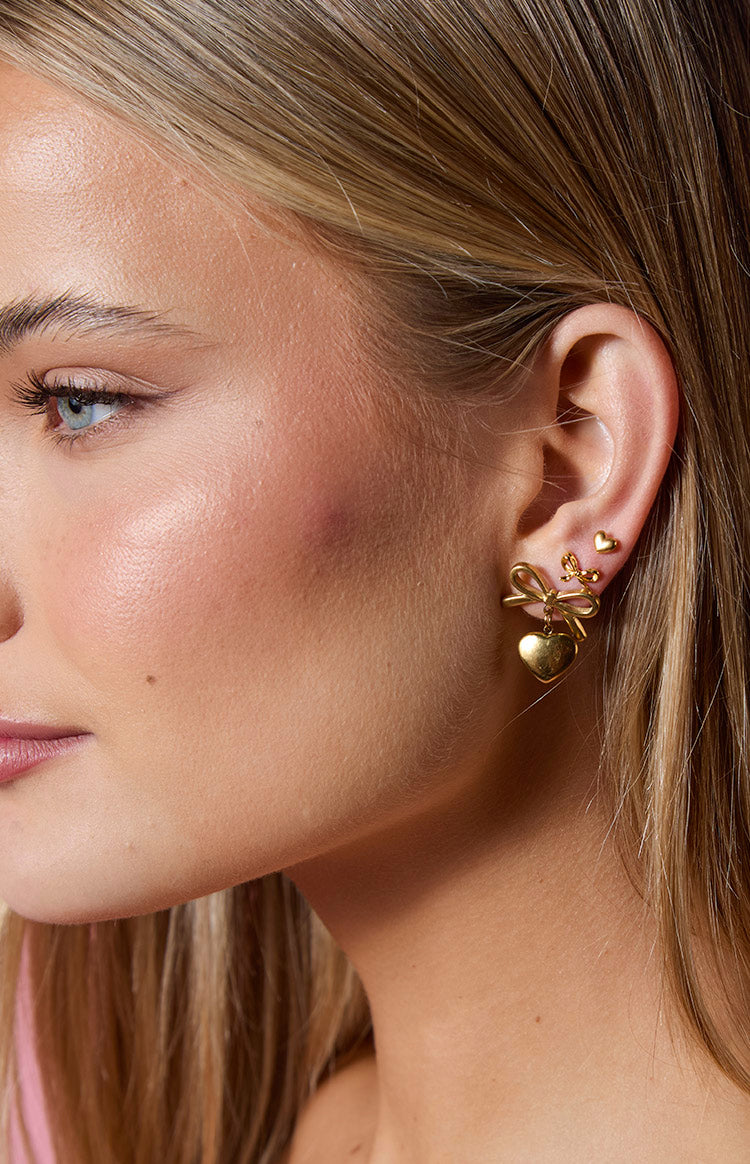 Close-up of three gold earrings: a bow with a dangling heart, a small heart stud, and a tiny ball stud