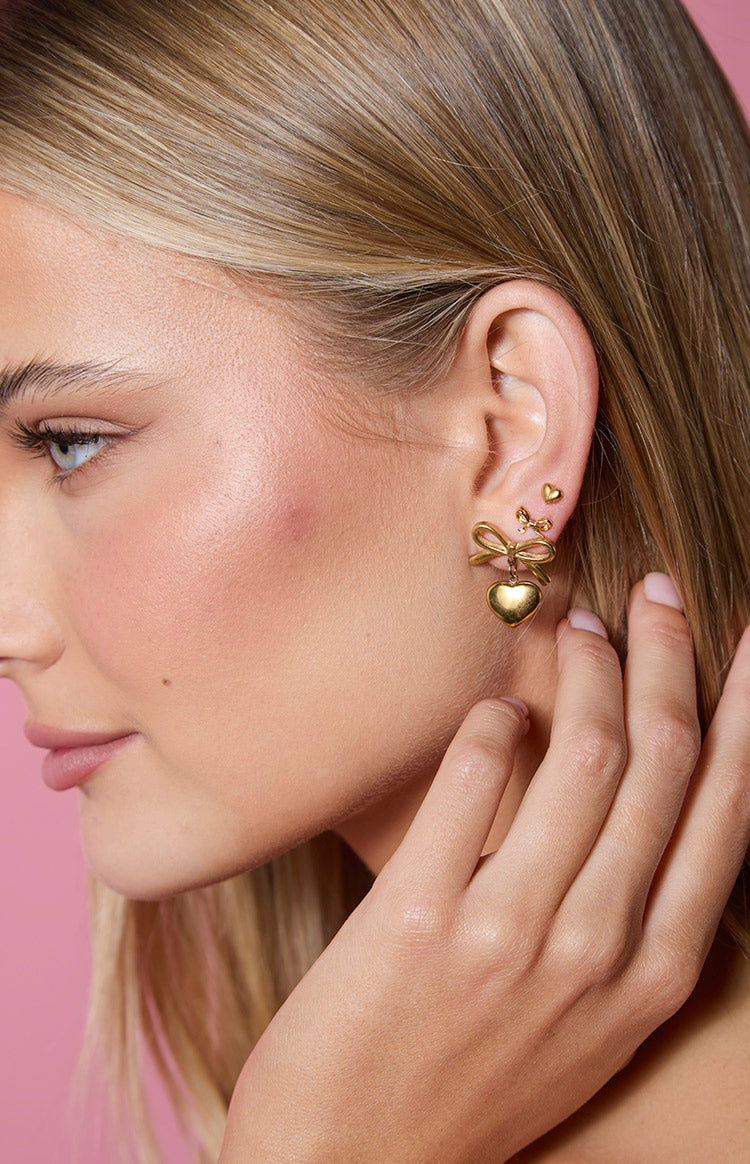 Close-up of three gold earrings: a small heart stud, a larger heart stud, and a bow with a dangling heart