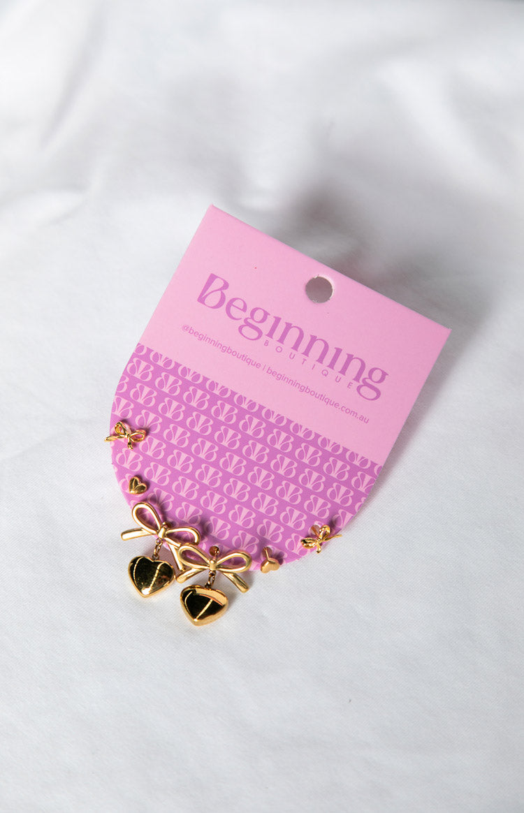 Three pairs of gold earrings including heart drop earrings with bows, small heart studs, and small bow studs