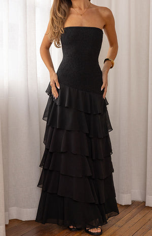 Our female model wearing a black strapless maxi dress with a shirred or smocked bodice and a tiered ruffled skirt.