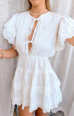 Close-up of the white mini dress showing pintuck bodice, lace-up V-neck, and eyelet lace trim on tiered skirt