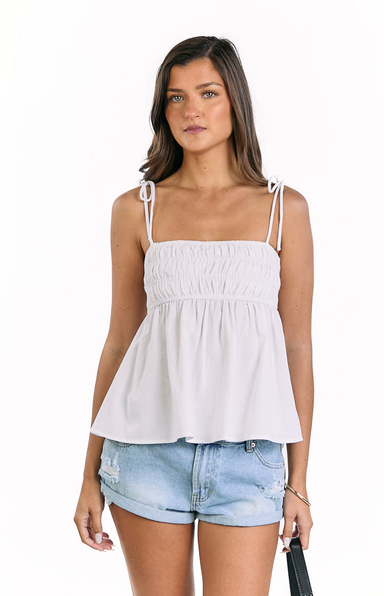Front view of a white ruched tie strap top with a square neckline and a flowy peplum hem