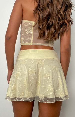 Rear view of a yellow mesh mini skirt with floral lace embroidery, a wide waistband, and a scalloped hem