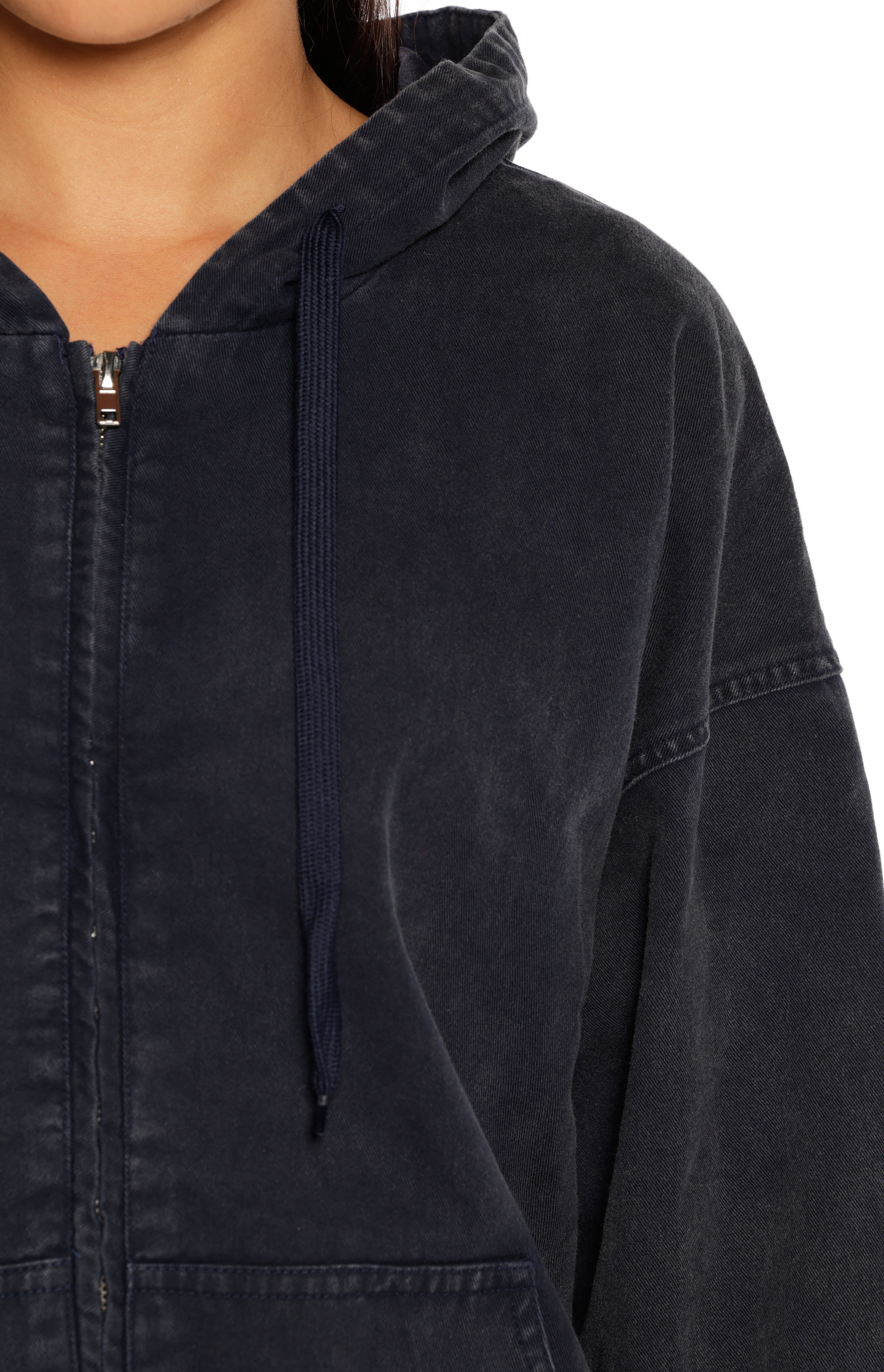 A close-up focusing on the upper chest and shoulder of an oversized dark washed zip-up hoodie or jacket in a deep navy or charcoal color. The garment has a slightly faded textured appearance a hood and navy blue drawstrings hanging from the neck. A silver zipper runs down the center and the jacket features dropped shoulders and a generous sleeve size.