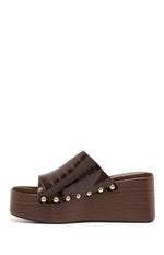 A side profile of a single dark brown platform wedge mule features a wide strap made of glossy crocodile-embossed faux leather. The strap is secured to the sole with a line of small gold-toned studs. The sole is a chunky platform wedge with a simulated dark wood-grain finish.