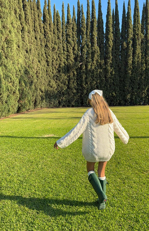 Girl showing the back of an oversized cream cable-knit sweater dress. The image highlights the cozy- thick knit pattern and long- roomy sleeves.