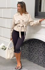 Cream cropped trench coat with double-breasted front, tie waist, wide lapels, shoulder epaulets, and cuff straps