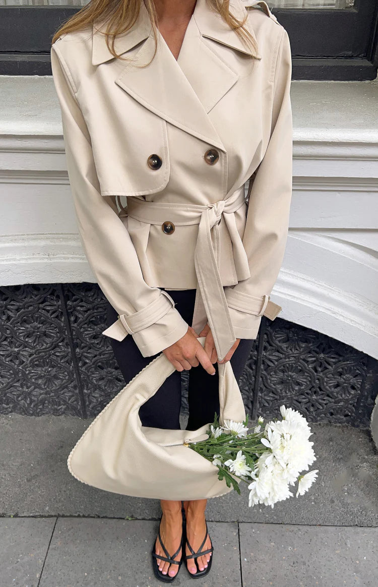 Cream cropped trench coat with double-breasted front, notched lapels, storm flap, shoulder epaulettes, and tied waist belt