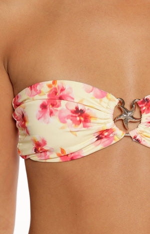 A tightly cropped image focuses on the center of a strapless bandeau bikini top. The swimwear fabric is a light yellow or cream color printed with large pink and red tropical flowers. The band is gathered and cinched in the middle by a silver-toned metal ring that holds a starfish charm.