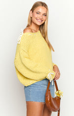 Side view of a chunky knit yellow sweater with white tie details on the shoulder and cuffs