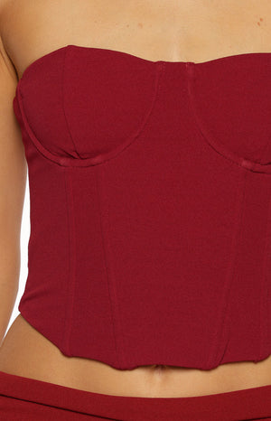 A close-up of our model highlights a strapless bustier top in a burgundy stretch fabric. This top features a sweetheart neckline and structured vertical boning resembling a corset with a pointed asymmetrical hemline at the waist.
