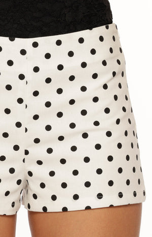 A close-up shot of our model wearing a pair of high-waisted shorts in a white fabric patterned with black polka dots. These shorts have a simple tailored cut.