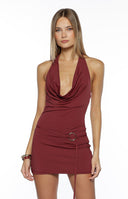 Our model wearing a halter mini dress in a deep burgundy or burnt red stretch fabric. This dress features a dramatic plunging cowl neckline that drapes low over the bust and a fitted mini skirt. The waist is cinched by a thin band of fabric that wraps around and is accented with a large silver circular ring and a dangling tie.