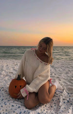 Cream knit oversized sweater with an off-shoulder neckline and pink ribbon details woven along the shoulder and cuffs