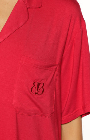 A close-up of our model wearing a two-piece pajama set in a vibrant solid red color. This top is a short-sleeve button-down shirt with a notched collar an oversized fit and a single breast pocket embroidered with a dark monogram (possibly a 'B'). The fabric appears to be a soft lightweight knit.