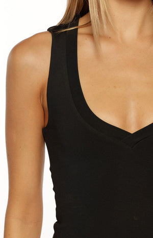 A close-up of our model highlights a fitted sleeveless tank top in solid black. This top features a deep U-neckline layered over a subtle V-neck inset creating a slight crossover effect at the bust and cropped with a thin white waistband visible beneath it.