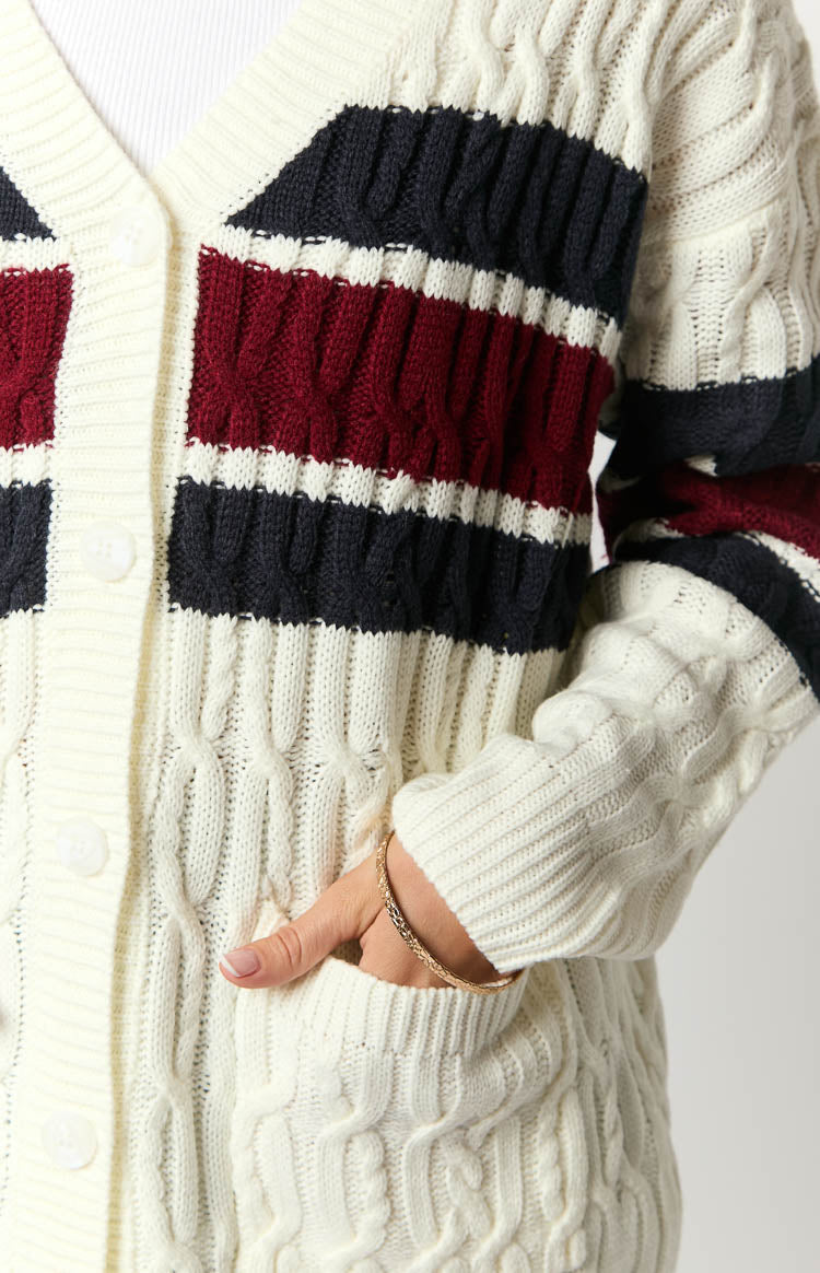 Close-up of the cream cable knit cardigan with navy and burgundy stripes, front buttons, and a visible pocket