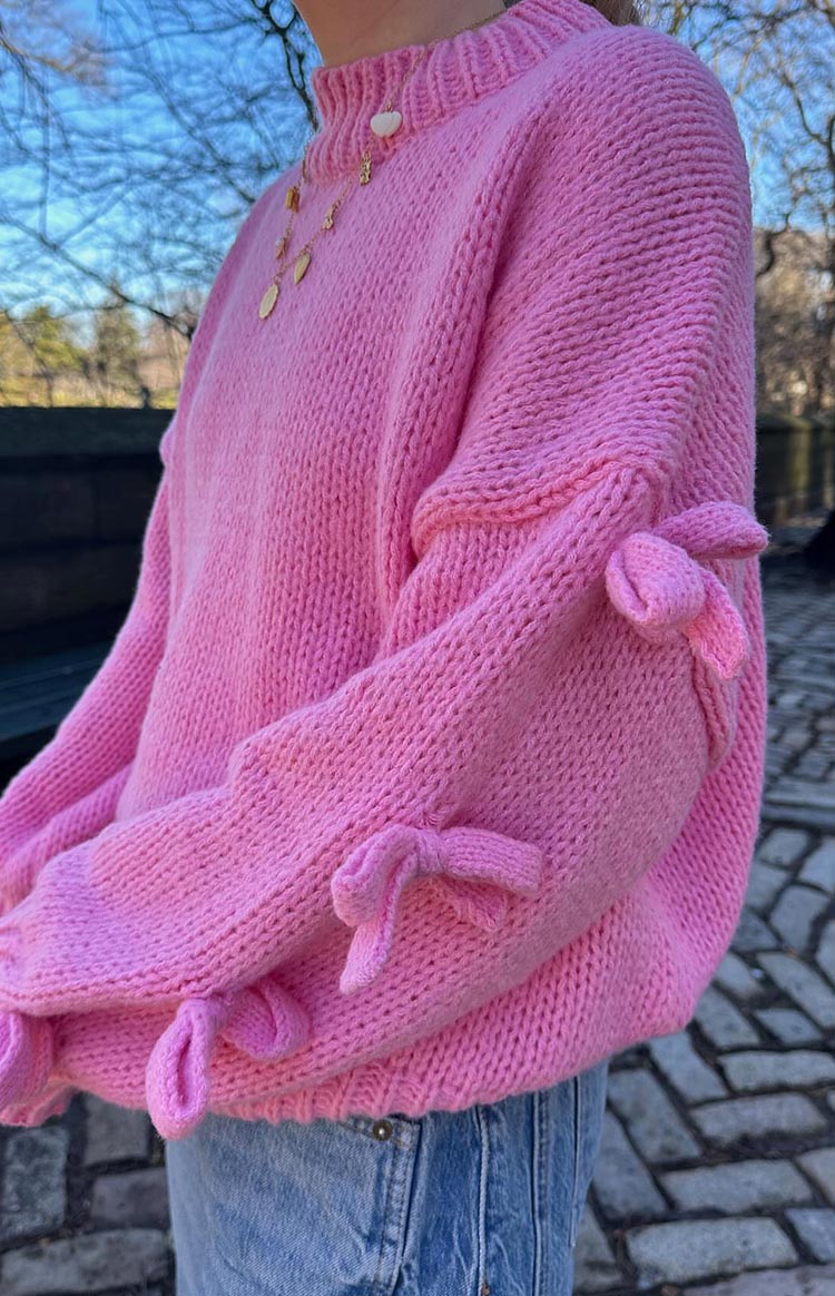 Close-up of a pink chunky knit jumper showing ribbed mock neck and multiple bow details on the sleeves