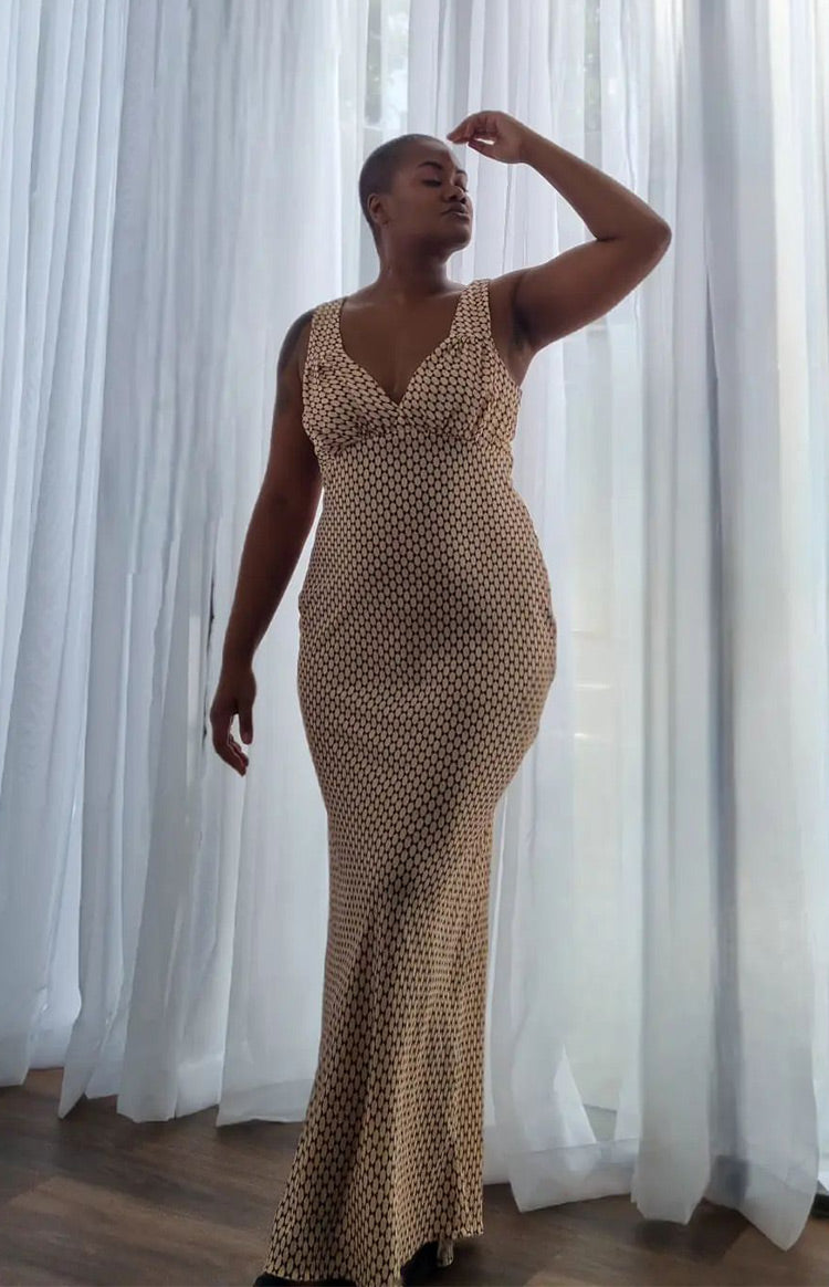 Full length view of a beige maxi dress with a black geometric pattern, V-neckline, and a fitted silhouette that flares at the hem